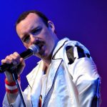 Freddie Mercury impersonator performing live in a white jacket with microphone.