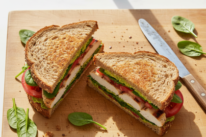 Grilled chicken sandwich on whole grain bread with spinach, tomato, and avocado on a wooden cutting board.